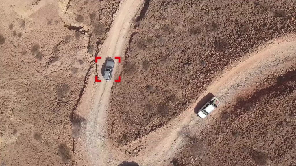 Overhead drone view of two pickup trucks on a desert track, one held in a red tracking reticle